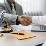 Cliente e representante jurídico apertando as mãos em escritório, com balança da justiça, martelo de juiz e notebook sobre a mesa, simbolizando acordo e antecipação de crédito judicial.